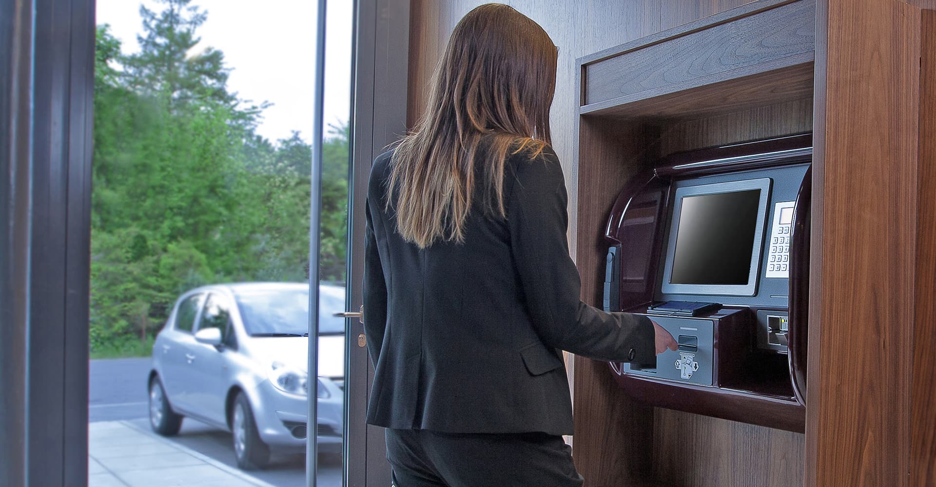 A woman uses a self check in terminal inside a building while a car is parked outside