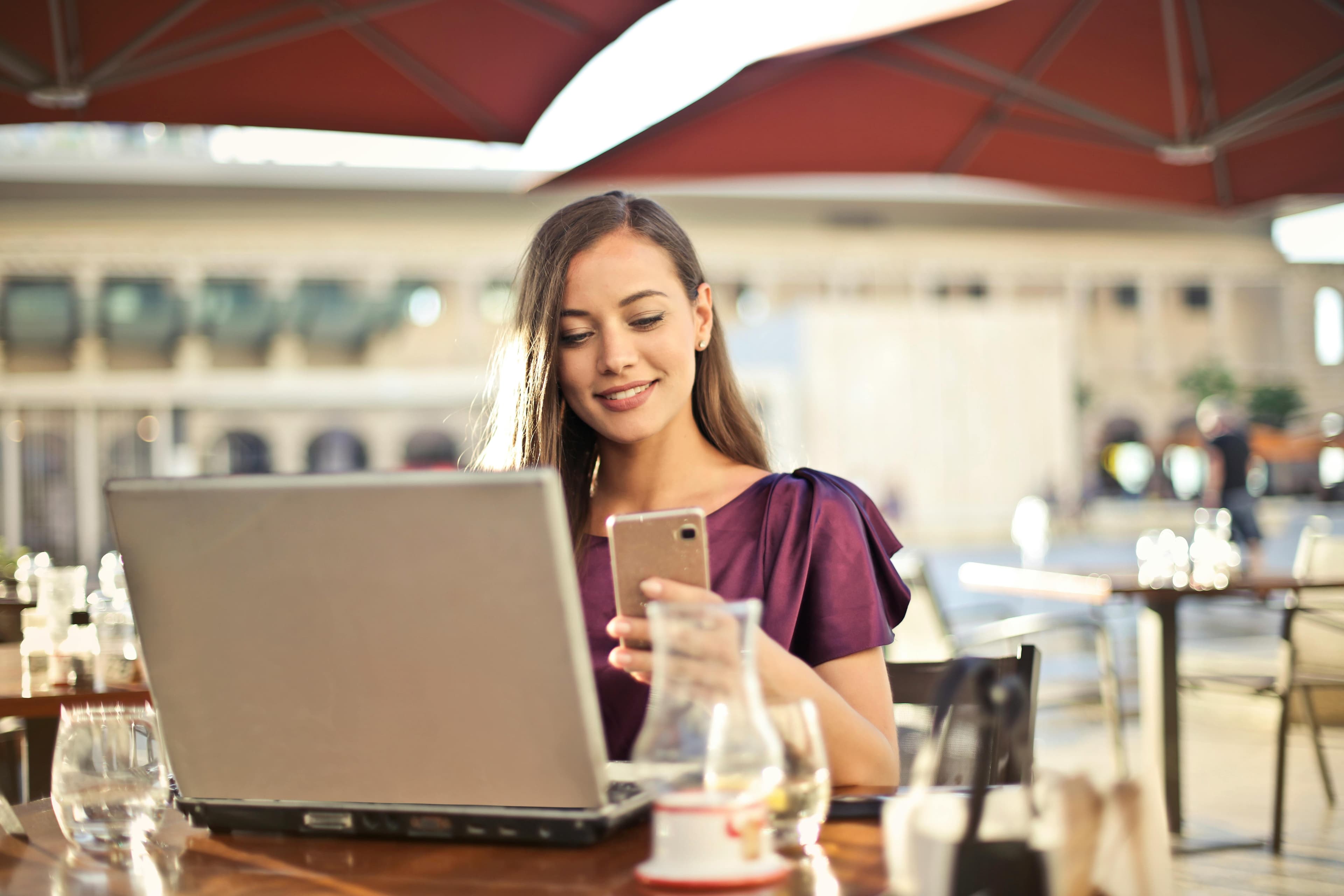 A woman sits at an outdoor cafe table and uses a smartphone while she sits in front of a laptop.
